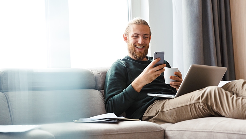 a bearded person sitting on the couch with their phone, coffee and a laptop
